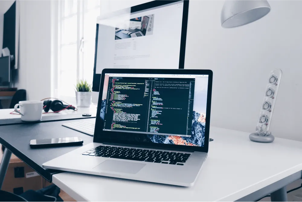 An office desk with a computer and open laptop with code on the screen.
